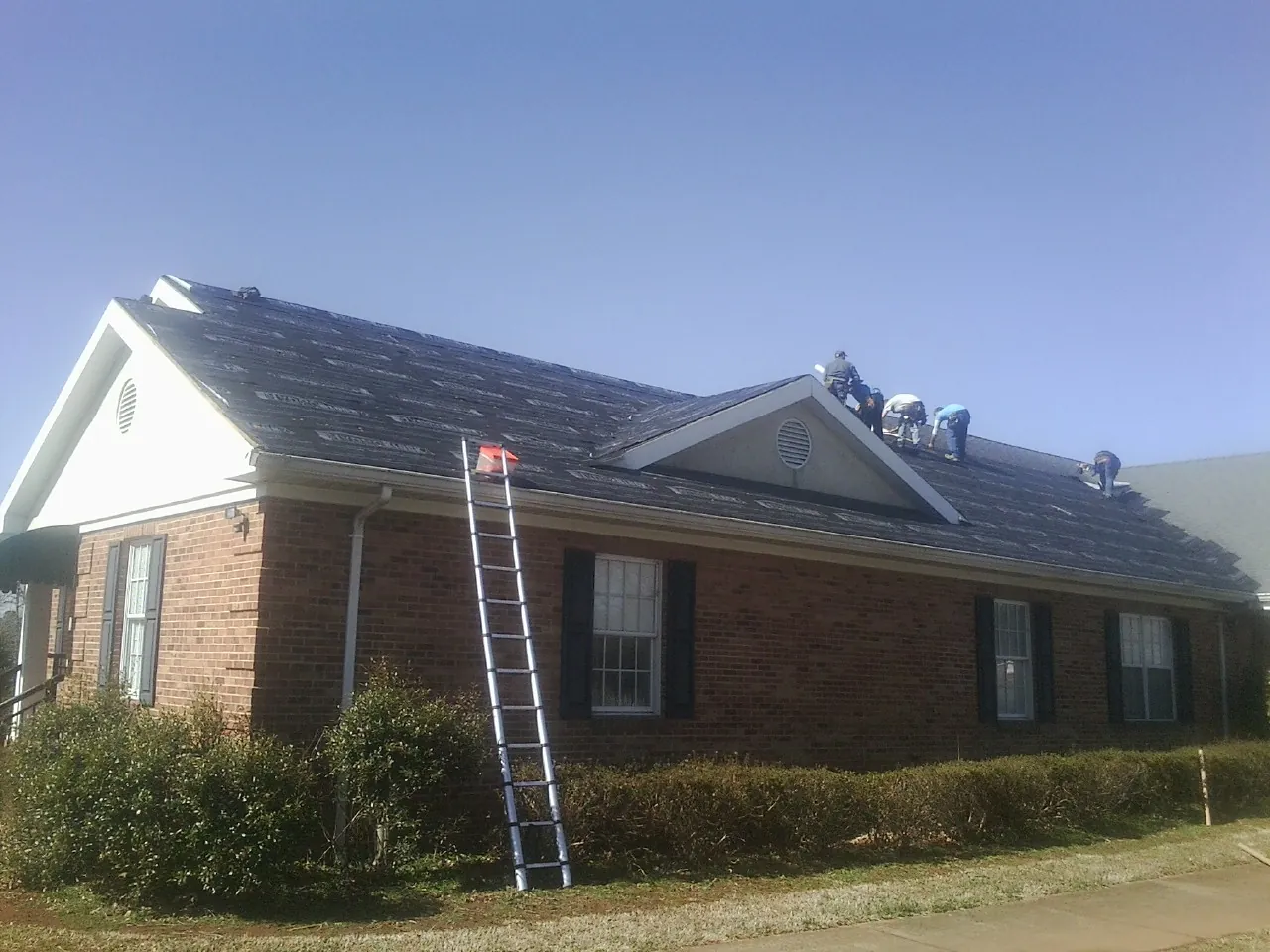 Crew tearing shingles off a brick ranch roof with a ladder set — Metal Roof Repair work by our crew in Emporia